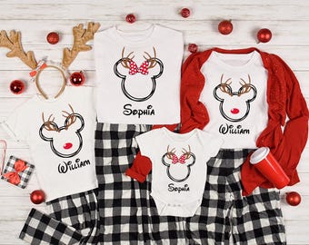 May include: A collection of white Christmas shirts and a onesie with a reindeer Mickey Mouse design. The shirts have the names "William" and "Sophia" printed on them. The shirts are paired with black and white plaid pajama pants and red accents.