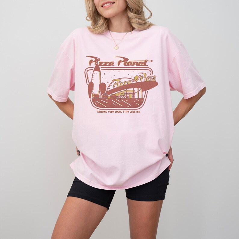 Puede incluir: Camiseta rosa claro con un dise&ntilde;o retro de Pizza Planet, que incluye un cohete y el texto "Serving Your Local Star Cluster". La camiseta tiene mangas cortas y un corte holgado.