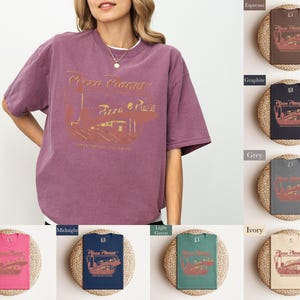 Puede incluir: Una camiseta de color malva con un gr&aacute;fico dorado de "Pizza Planet" de estilo vintage. La imagen muestra la camiseta en una persona y tambi&eacute;n muestra la camiseta en varios colores, incluyendo espresso, grafito, gris e marfil.