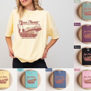Puede incluir: Una camiseta amarilla clara con el logo de Pizza Planet en marr&oacute;n y rojo. El logo presenta un cohete y un restaurante tem&aacute;tico espacial. El texto "Serving Your Local Star Blaster" est&aacute; debajo del logo. La camiseta la lleva una persona.