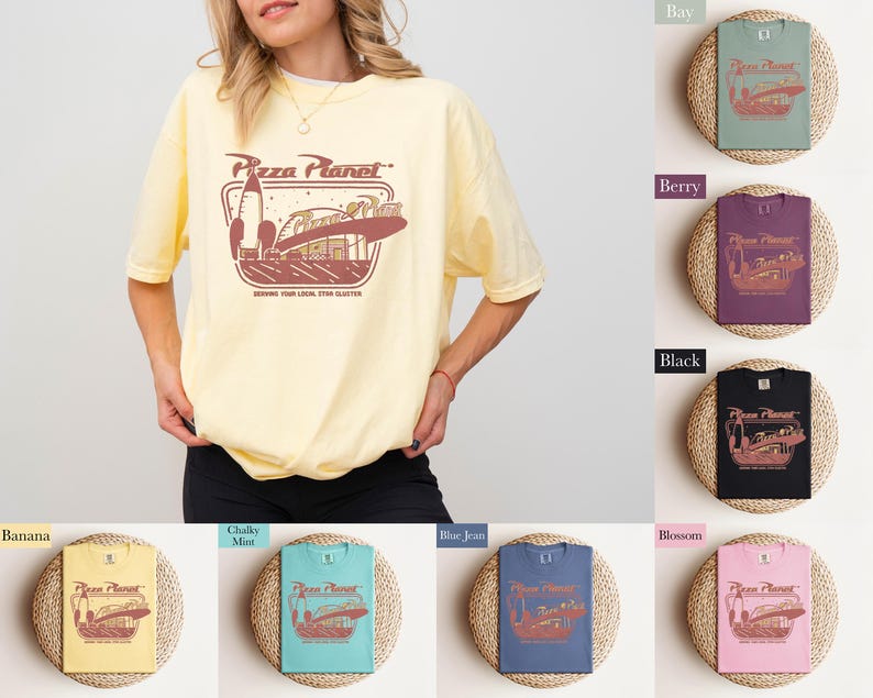 Puede incluir: Una camiseta amarilla clara con el logo de Pizza Planet en marr&oacute;n y rojo. El logo presenta un cohete y un restaurante tem&aacute;tico espacial. El texto "Serving Your Local Star Blaster" est&aacute; debajo del logo. La camiseta la lleva una persona.