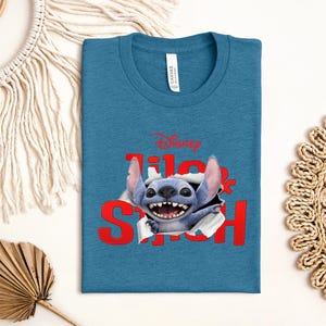 Puede incluir: Camiseta azul turquesa con un gr&aacute;fico de Stitch de Lilo & Stitch de Disney. El texto rojo dice "Lilo & Stitch" con Stitch rompiendo las letras. La camiseta tiene cuello redondo y una etiqueta que dice "Canvas".