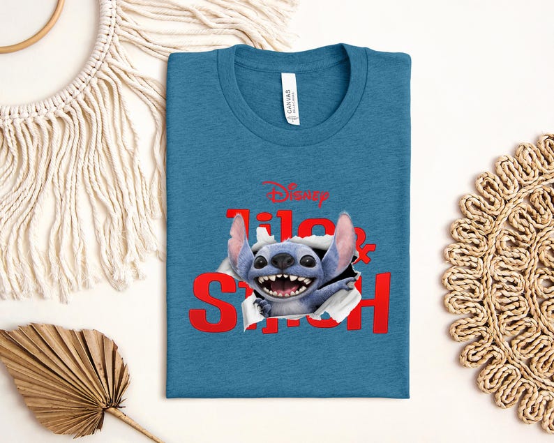 Puede incluir: Camiseta azul turquesa con un gr&aacute;fico de Stitch de Lilo & Stitch de Disney. El texto rojo dice "Lilo & Stitch" con Stitch rompiendo las letras. La camiseta tiene cuello redondo y una etiqueta que dice "Canvas".
