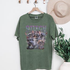 K&ouml;nnte beinhalten: Gr&uuml;nes T-Shirt mit einer Grafik der Figur Stitch aus dem Film Lilo & Stitch. Das Wort "STITCH" ist in einer lila, glitzernden Schrift &uuml;ber der Figur gedruckt. Das Bild enth&auml;lt mehrere Darstellungen von Stitch.