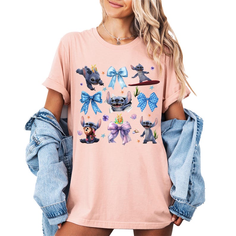Puede incluir: Una camiseta rosa claro con un dise&ntilde;o de personajes de dibujos animados y lazos coloridos. Los personajes son azules con orejas grandes y est&aacute;n en varias poses. Los lazos son en tonos azules y morados.