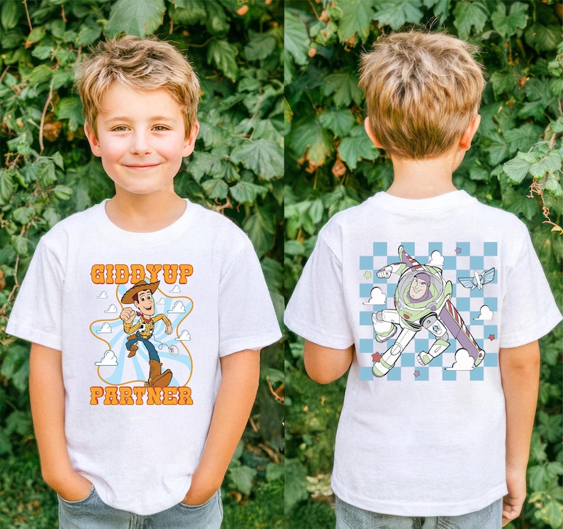 May include: White t-shirt featuring a cartoon graphic of Woody from Toy Story with the text "Giddyup Partner". The back of the shirt has a graphic of Buzz Lightyear. The shirt is worn by a young child.