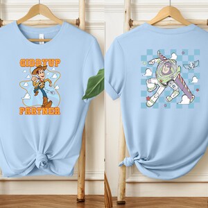 May include: Light blue t-shirts with cartoon graphics. One shirt features Woody with the text "Giddy Up Partner". The other shirt displays Buzz Lightyear. Both shirts are displayed on wooden hangers against a light background.