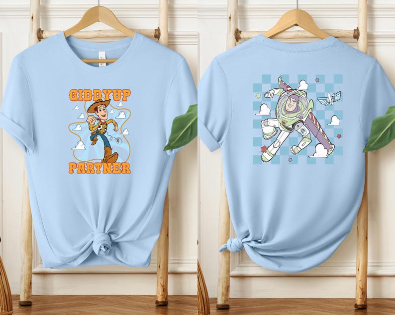 May include: Light blue t-shirts with cartoon graphics. One shirt features Woody with the text "Giddy Up Partner". The other shirt displays Buzz Lightyear. Both shirts are displayed on wooden hangers against a light background.