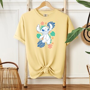 Puede incluir: Una camiseta amarillo claro con un personaje de dibujos animados en azul, blanco y verde, rodeado de motivos florales y de hojas. La camiseta est&aacute; anudada a la cintura y colgada en un perchero de madera.