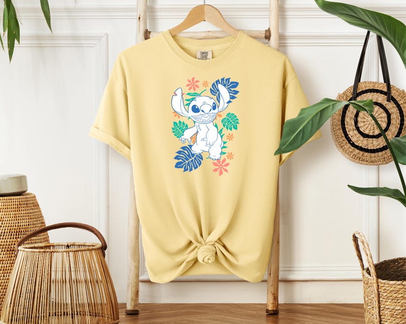 Puede incluir: Una camiseta amarillo claro con un personaje de dibujos animados en azul, blanco y verde, rodeado de motivos florales y de hojas. La camiseta est&aacute; anudada a la cintura y colgada en un perchero de madera.