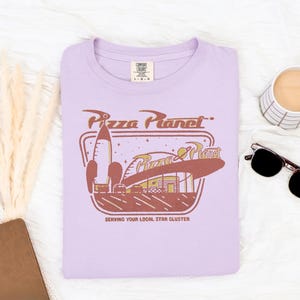 Puede incluir: Una camiseta lila con el logo de Pizza Planet en marr&oacute;n y dorado. El dise&ntilde;o incluye un cohete, las palabras "Pizza Planet" y el lema "Serving Your Local Star Cluster". La camiseta es de la marca Comfort Colors.