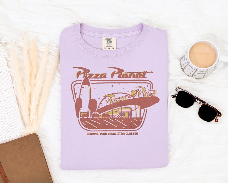Puede incluir: Una camiseta lila con el logo de Pizza Planet en marr&oacute;n y dorado. El dise&ntilde;o incluye un cohete, las palabras "Pizza Planet" y el lema "Serving Your Local Star Cluster". La camiseta es de la marca Comfort Colors.