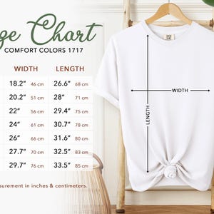 K&ouml;nnte beinhalten: Ein wei&szlig;es T-Shirt h&auml;ngt an einem Holzb&uuml;gel, mit einer Gr&ouml;&szlig;entabelle links. Die Tabelle zeigt Gr&ouml;&szlig;en von S bis 4XL, mit Ma&szlig;en in Zoll und Zentimetern. Die Tabelle enth&auml;lt Breiten- und L&auml;ngenangaben f&uuml;r jede Gr&ouml;&szlig;e.