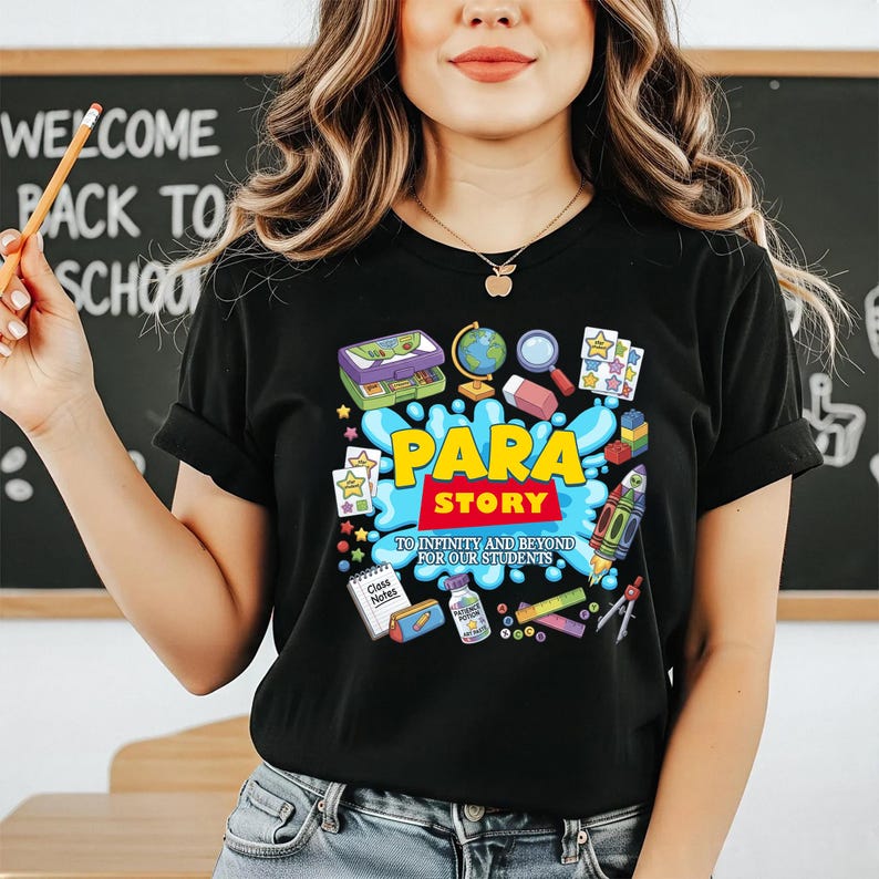 K&ouml;nnte beinhalten: Schwarzes T-Shirt mit einem farbenfrohen Grafikdesign. Das Design enth&auml;lt die Worte "PARA STORY" und den Satz "TO INFINITY AND BEYOND FOR OUR STUDENTS." Das Design beinhaltet schulbezogene Illustrationen wie Globus, Rakete und Sterne.