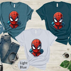 May include: Three t-shirts in teal and light blue with a cartoon Spider-Man graphic. The shirts are displayed with a pair of blue jeans, black and white sneakers, and headphones. The text on the image reads "Heather Deep Teal", "Deep Teal", and "Light Blue".
