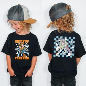 May include: Black t-shirt featuring a cartoon graphic of Woody with the text "Giddyup Partner" on the front. The back of the shirt has a graphic of Buzz Lightyear. The child is wearing a gray and tan trucker hat.