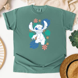 Puede incluir: Una camiseta de color verde azulado con un personaje de dibujos animados blanco, orejas grandes y una amplia sonrisa, rodeado de dise&ntilde;os florales y de hojas coloridas. Prenda de vestir informal.