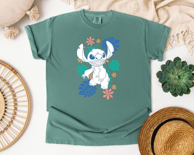 Puede incluir: Una camiseta de color verde azulado con un personaje de dibujos animados blanco, orejas grandes y una amplia sonrisa, rodeado de dise&ntilde;os florales y de hojas coloridas. Prenda de vestir informal.