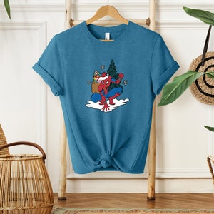 Puede incluir: Camiseta de color turquesa con un dibujo animado de Spider-Man con un gorro de Pap&aacute; Noel, sosteniendo regalos y un &aacute;rbol de Navidad. El dise&ntilde;o incluye copos de nieve y una escena nevada. La camiseta est&aacute; colgada en una percha de madera.