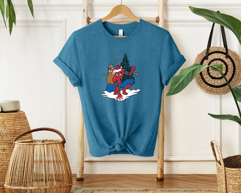Puede incluir: Camiseta de color turquesa con un dibujo animado de Spider-Man con un gorro de Pap&aacute; Noel, sosteniendo regalos y un &aacute;rbol de Navidad. El dise&ntilde;o incluye copos de nieve y una escena nevada. La camiseta est&aacute; colgada en una percha de madera.
