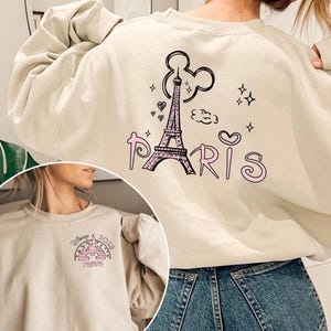 May include: Beige sweatshirt with a graphic of the Eiffel Tower, Mickey Mouse ears, hearts, and the word "Paris" in pink. The sweatshirt also features a smaller graphic of a castle with the text "Disney 2024 Mom".
