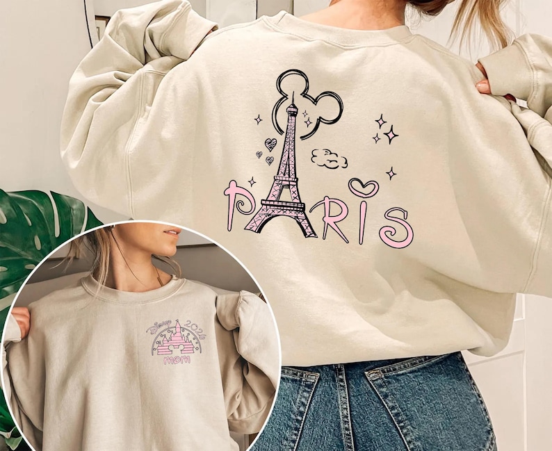 May include: Beige sweatshirt with a graphic of the Eiffel Tower, Mickey Mouse ears, hearts, and the word "Paris" in pink. The sweatshirt also features a smaller graphic of a castle with the text "Disney 2024 Mom".