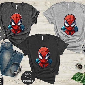 May include: Three t-shirts featuring a cartoon Spider-Man design in red, blue, and white. The shirts are dark gray, athletic heather gray, and dark gray heather. The image includes a pair of blue jeans, headphones, and a cup of coffee.