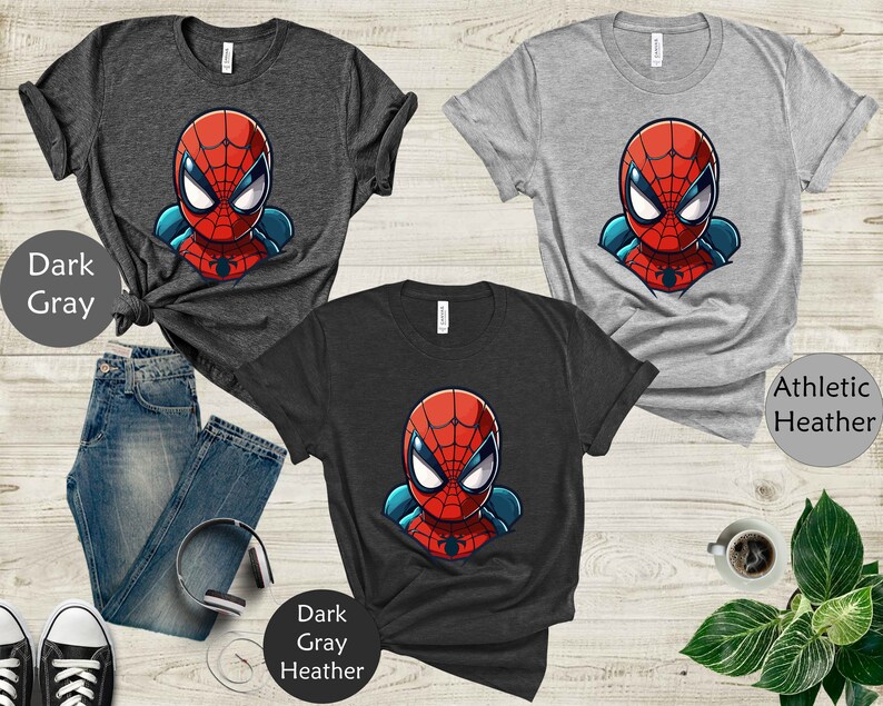 May include: Three t-shirts featuring a cartoon Spider-Man design in red, blue, and white. The shirts are dark gray, athletic heather gray, and dark gray heather. The image includes a pair of blue jeans, headphones, and a cup of coffee.