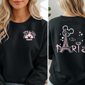 May include: Black sweatshirt with pink graphic designs. The front features a castle design with the text "Disney 2026 mom". The back displays the Eiffel Tower, Mickey Mouse ears, and the word "Paris".