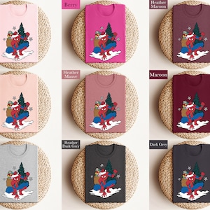 Puede incluir: Una colecci&oacute;n de camisetas en varios colores, incluyendo rosa, baya y gris, cada una con un dise&ntilde;o de superh&eacute;roe con gorro de Pap&aacute; Noel, &aacute;rbol de Navidad y regalos. Presentadas sobre esteras tejidas.