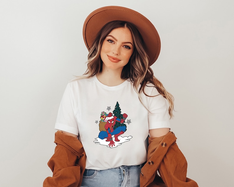 Puede incluir: Camiseta blanca con un gr&aacute;fico festivo de Spider-Man con un gorro de Pap&aacute; Noel, llevando un &aacute;rbol de Navidad y regalos. El superh&eacute;roe est&aacute; representado con su traje rojo y azul. El dise&ntilde;o incluye una bolsa de regalos y estrellas.