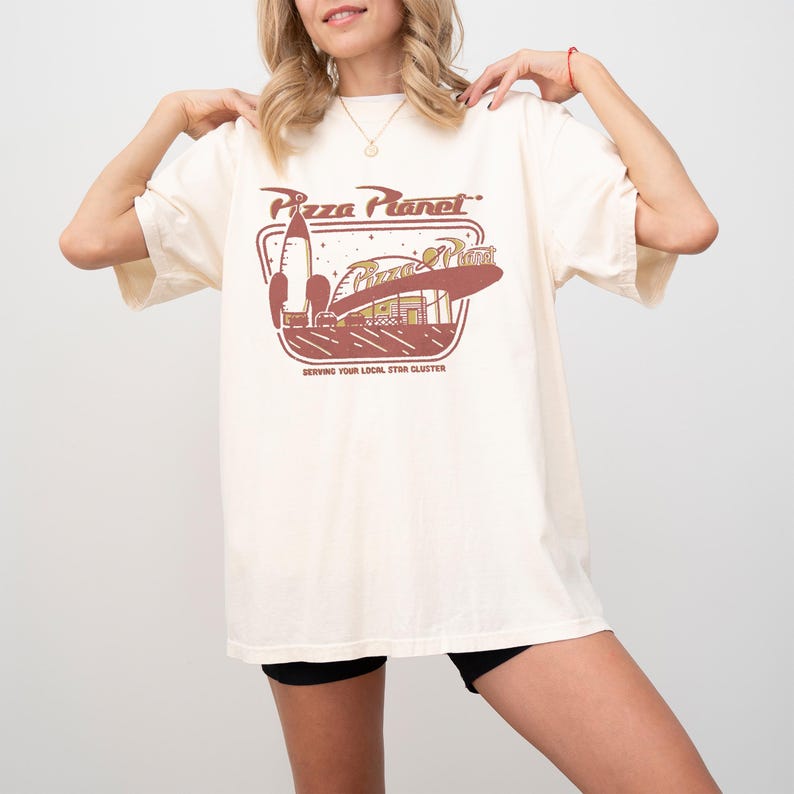 Puede incluir: Camiseta color crema con un gr&aacute;fico de Pizza Planet. El dise&ntilde;o incluye un cohete, un restaurante y el texto "Pizza Planet" y "Serving Your Local Star Cluster" en marr&oacute;n. La camiseta tiene mangas cortas y un corte holgado.