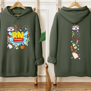 K&ouml;nnte beinhalten: Ein olivgr&uuml;ner Kapuzenpullover mit einem Front-Grafik-Design mit dem Text "RN STORY" in Gelb und Rot, umgeben von medizinischen Illustrationen. Die R&uuml;ckseite des Hoodies hat ein vertikales Design mit medizinischen Symbolen.
