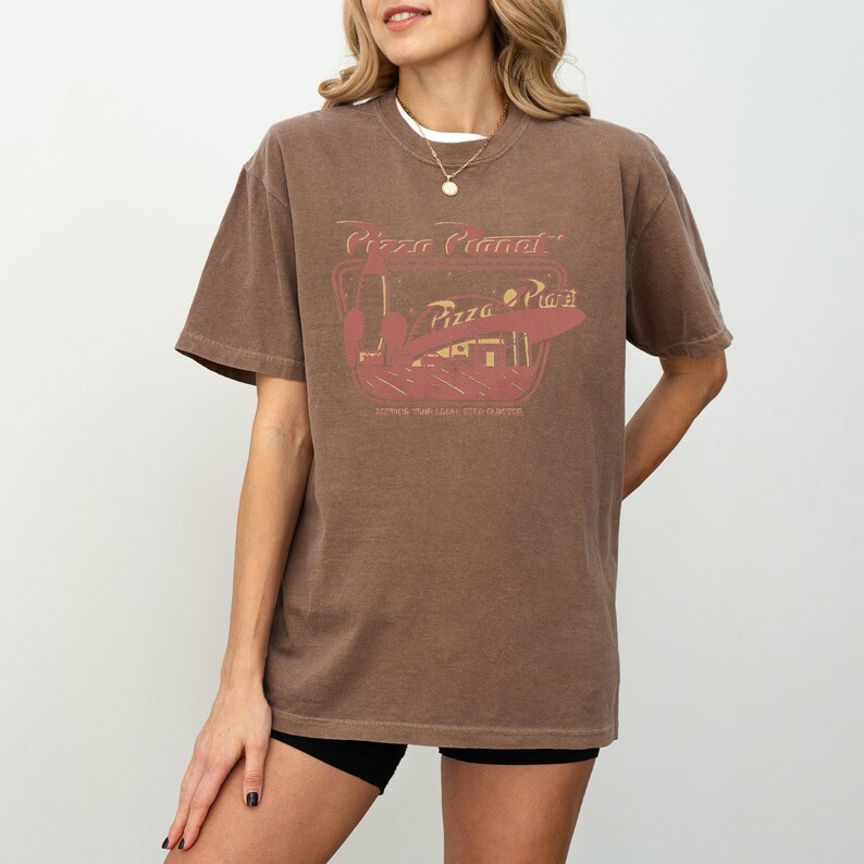 Puede incluir: Camiseta marr&oacute;n con un dise&ntilde;o de Pizza Planet en rojo y dorado. La camiseta tiene un aspecto vintage y cuello redondo. El dise&ntilde;o incluye el texto "Pizza Planet" y "Pizza Place".
