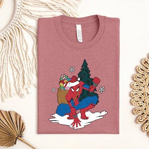 Puede incluir: Camiseta de color malva con un dise&ntilde;o de Spider-Man con gorro de Pap&aacute; Noel, arrodillado sobre una nube con un saco de regalos y un &aacute;rbol de Navidad. Copos de nieve blancos salpican el dise&ntilde;o. La camiseta tiene cuello redondo y una etiqueta "Canvas".