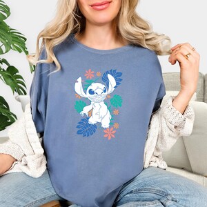 Puede incluir: Camiseta azul con un personaje de dibujos animados blanco, orejas grandes y una amplia sonrisa, rodeado de dise&ntilde;os florales y de hojas coloridas. La camiseta es de color azul liso.