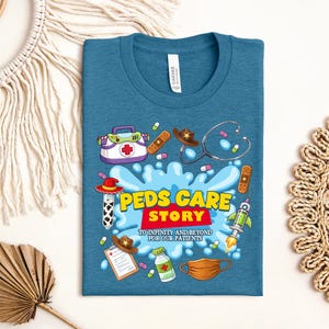 May include: Teal t-shirt with a colorful graphic design. The design includes medical-themed illustrations such as a stethoscope, pills, and a first aid kit. Text on the shirt reads "Peds Care Story" and "To Infinity and Beyond for Our Patients."