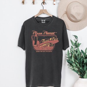 Puede incluir: Camiseta gris oscuro con un gr&aacute;fico de Pizza Planet, que incluye un cohete y el texto "Pizza Planet". La camiseta est&aacute; colgada en una percha de madera. Un sombrero beige es visible en el fondo.