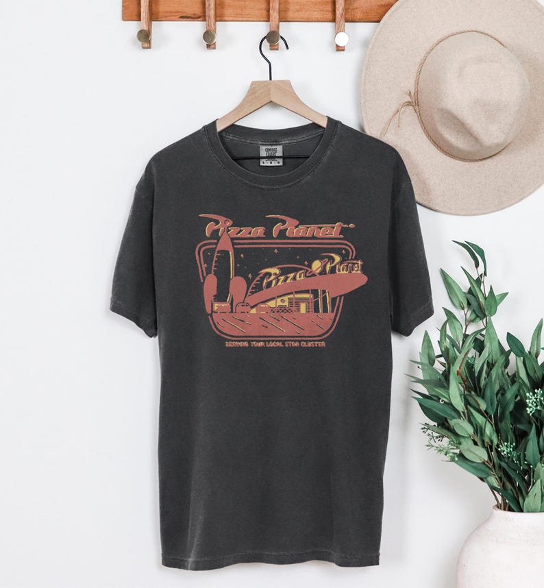Puede incluir: Camiseta gris oscuro con un gr&aacute;fico de Pizza Planet, que incluye un cohete y el texto "Pizza Planet". La camiseta est&aacute; colgada en una percha de madera. Un sombrero beige es visible en el fondo.