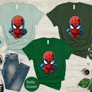 May include: Three t-shirts in mint, heather forest, and kelly green, each featuring a cartoon Spider-Man graphic. The shirts are displayed with a pair of blue jeans, black sneakers, and other accessories on a wooden surface.