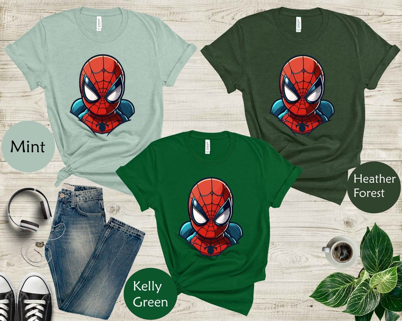 May include: Three t-shirts in mint, heather forest, and kelly green, each featuring a cartoon Spider-Man graphic. The shirts are displayed with a pair of blue jeans, black sneakers, and other accessories on a wooden surface.