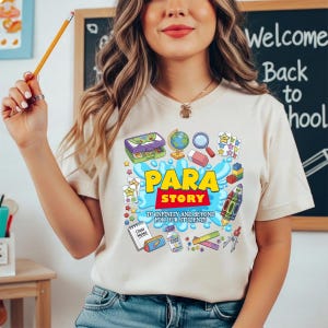 K&ouml;nnte beinhalten: Beigefarbenes T-Shirt mit einem farbenfrohen Grafikdesign. Das Design zeigt die Worte "PARA STORY" und Illustrationen zum Thema Schule, wie einen Globus, eine Lupe und eine Rakete. Der Text "TO INFINITY AND BEYOND FOR OUR STUDENTS" ist ebenfalls vorhanden.
