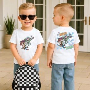 Puede incluir: Camiseta blanca con un gr&aacute;fico de Buzz Lightyear y Woody de Toy Story. La camiseta tiene la frase "To Infinity and Beyond!" en la espalda. El ni&ntilde;o lleva vaqueros azules y sostiene una mochila a cuadros blancos y negros.