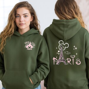 May include: Olive green hoodie with a front pocket and a hood. The front features a pink and white graphic with the text "Disney 2026 mom". The back displays a pink Eiffel Tower graphic with the word "Paris".