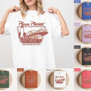 Puede incluir: Camiseta blanca con un gr&aacute;fico de Pizza Planet en marr&oacute;n y rojo. El dise&ntilde;o incluye un cohete, un edificio y el texto "Pizza Planet" y "Serving Your Local Star Cluster". Se muestran camisetas adicionales en varios colores.
