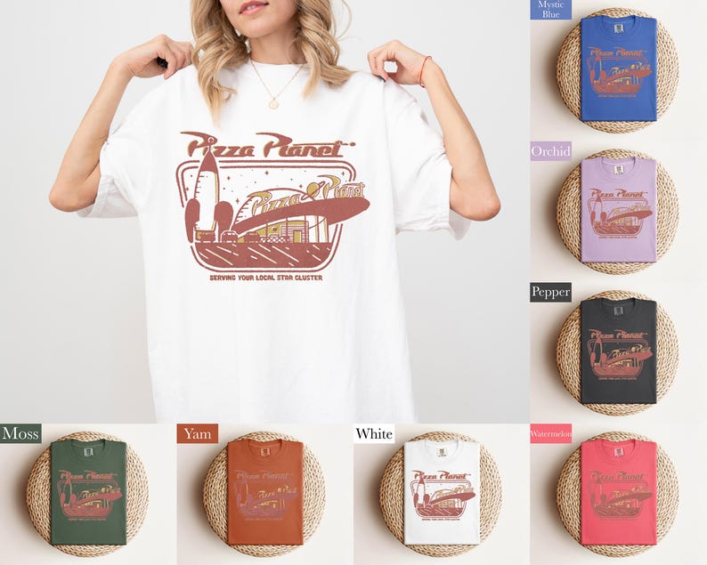 Puede incluir: Camiseta blanca con un gr&aacute;fico de Pizza Planet en marr&oacute;n y rojo. El dise&ntilde;o incluye un cohete, un edificio y el texto "Pizza Planet" y "Serving Your Local Star Cluster". Se muestran camisetas adicionales en varios colores.