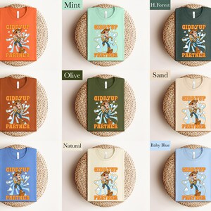 May include: A collection of t-shirts in various colors, each featuring a cartoon cowboy graphic with the text "GIDDYUP PARTNER". Colors include orange, mint, forest green, autumn, olive, sand, blue, natural, and baby blue.