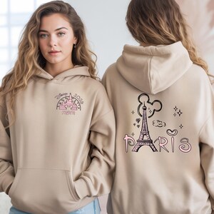 May include: Tan hoodie with a front pocket and a hood. The front features a pink and black graphic of a castle and the word "MOM". The back has a black Eiffel Tower graphic with the word "PARIS" in pink.