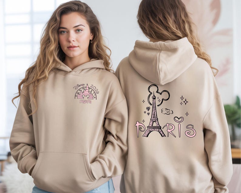 May include: Tan hoodie with a front pocket and a hood. The front features a pink and black graphic of a castle and the word "MOM". The back has a black Eiffel Tower graphic with the word "PARIS" in pink.