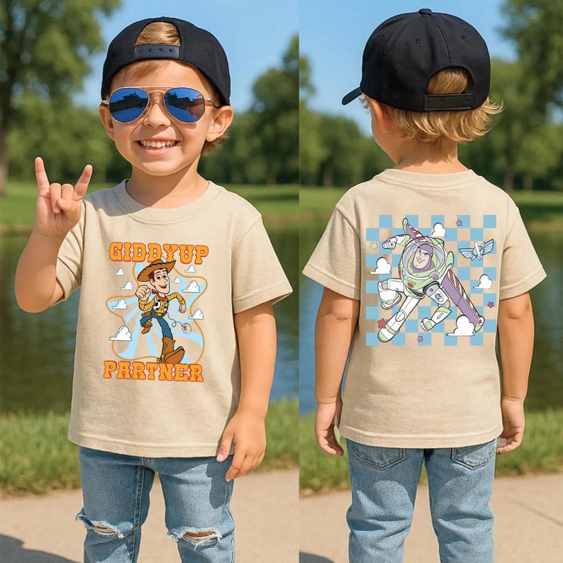 May include: A tan t-shirt featuring a cartoon of Woody with the text "Giddyup Partner" on the front. The back of the shirt has a cartoon of Buzz Lightyear. The child is wearing blue jeans and a black baseball cap.